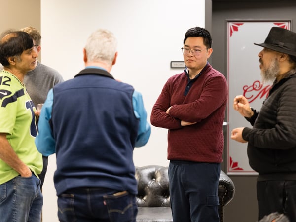A group of five men are standing in a room, engaged in a conversation. One man wears a bright green sports jersey, while another wears a maroon sweater and glasses.