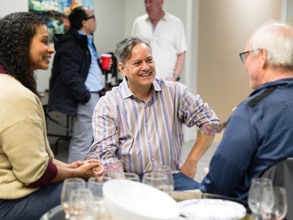 A group of people are engaged in conversation at a social gathering indoors, with a man in a striped shirt smiling warmly at his companions.