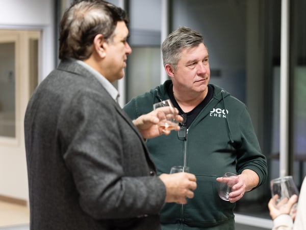 Two men are engaged in conversation during a social gathering in an indoor setting, both holding glasses of drinks.