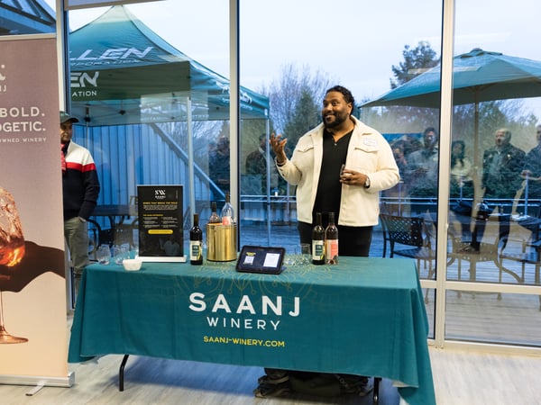A man stands behind a promotional table for Saanj Winery, gesturing while speaking during what appears to be a wine tasting or event.