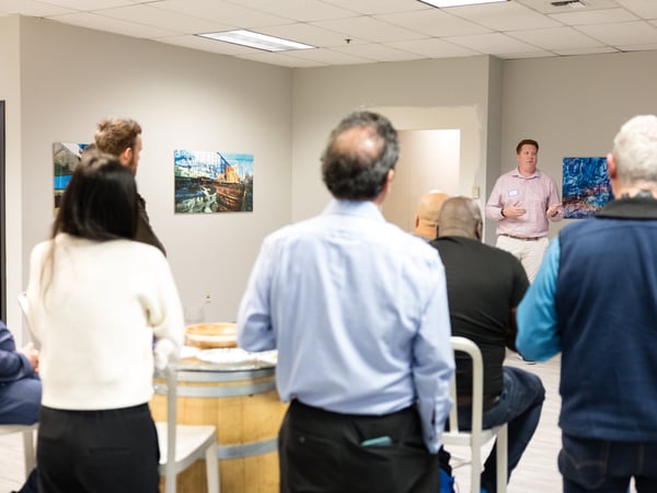 A man in a pink shirt is giving a presentation or talk to a group of people in an indoor space decorated with art.
