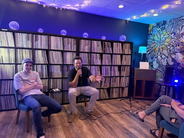 A photograph of a live discussion or podcast recording taking place indoors, featuring two men seated in front of a large wall of vinyl records while one speaks into a microphone.