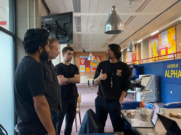 A group of four men are standing in a modern office or coworking space, engaged in a discussion. One man is gesturing with a thumbs-up while speaking to the others, who are listening attentively near a table with a laptop and snacks.