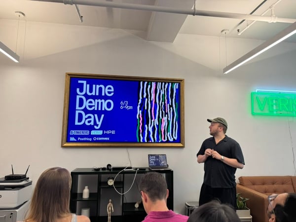 A photograph of a presenter wearing a cap and black shirt standing next to a large screen displaying 'June Demo Day' in front of an audience.