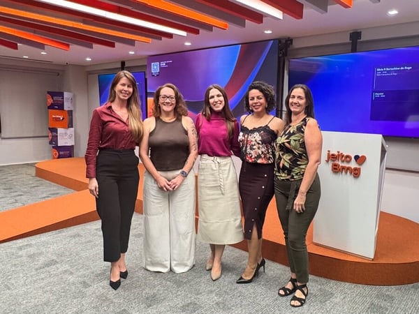 A group of five women posing for a photo in a modern office or event space featuring an orange stage and digital screens.