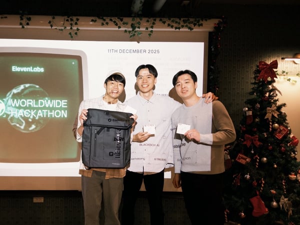 Three young men, presumably winners, pose for a photo in front of a projection screen displaying the logo and title for the "ElevenLabs WORLDWIDE HACKATHON," holding a backpack and cards as prizes in an indoor space decorated with Christmas garlands and a Christmas tree.