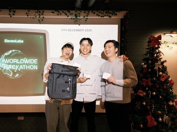 Three smiling men stand in front of a projection screen displaying the event title "ElevenLabs WORLDWIDE HACKATHON" and the date, holding prizes (a backpack and cards) at an indoor event decorated with Christmas greenery and a tree.