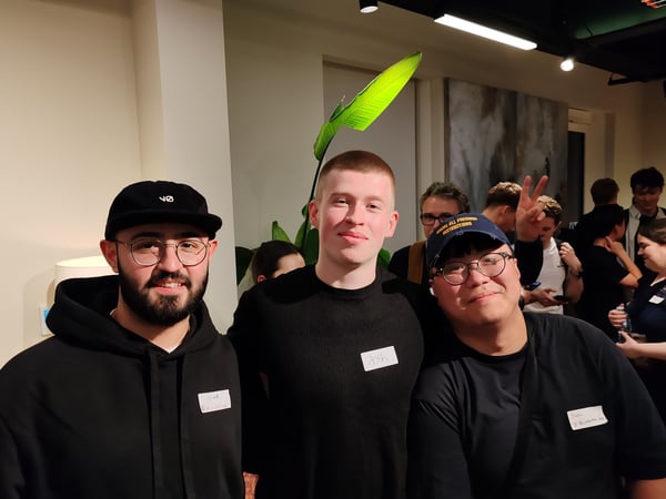A group portrait featuring three young men smiling at the camera in the foreground, taken at an indoor social or networking event with many other attendees visible in the warm, dimly lit background.