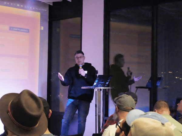 A man wearing a dark hoodie and jeans is giving a presentation, holding a microphone, in front of a projected screen displaying a user interface and large windows overlooking a city view at night. Several audience members are visible in the foreground, seen from behind.