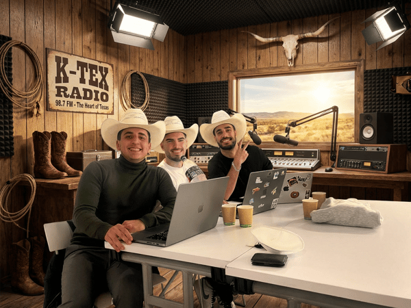 Three young men wearing cowboy hats are seated at a table in a rustic, wood-paneled radio studio, smiling at the camera while recording a show. The studio, decorated with Western artifacts like a longhorn skull and cowboy boots, features professional audio equipment and laptops.