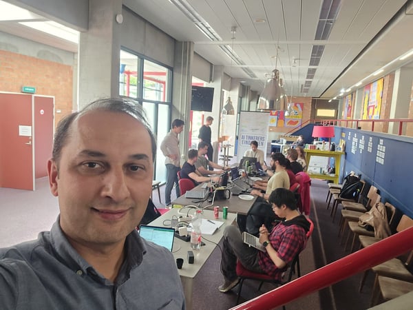 A selfie of a man in the foreground with a group of people working on laptops at a long table in a modern indoor workspace.