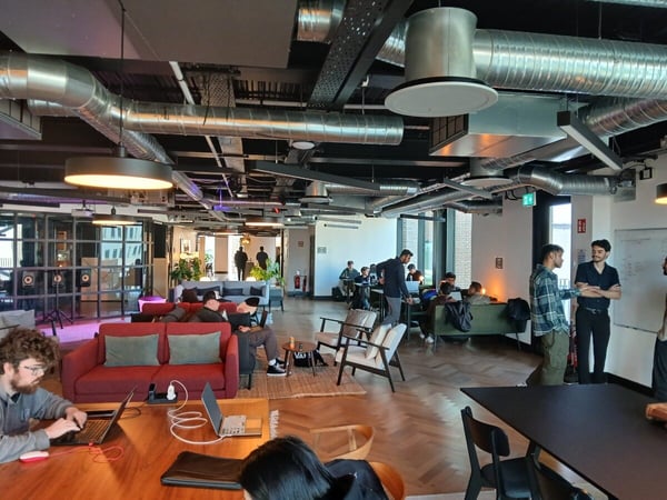 A wide-angle indoor shot of a modern, industrial-style co-working space featuring exposed ductwork, people working on laptops, and groups engaged in conversation.