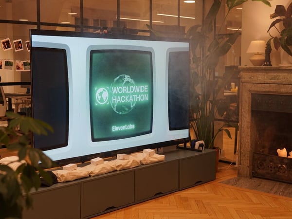 A large television screen displaying the title 'WORLDWIDE HACKATHON' in a retro green CRT style is set up in a warm, indoor office lounge area featuring a marble fireplace and large potted plants.