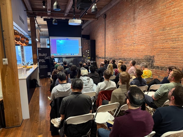 A group of people are seated in a room with exposed brick walls, attending a presentation shown on a large projector screen.