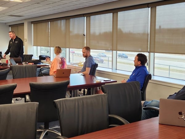 A group of people are gathered in a modern conference room for a meeting or presentation, with one person standing and leading the discussion.
