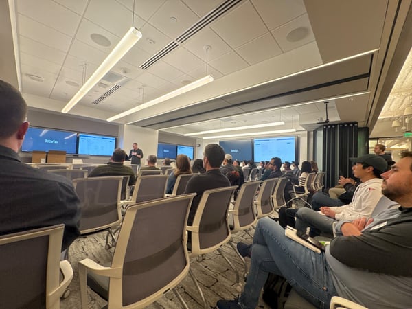 A photograph captures a large audience seated in a modern, well-lit conference room, listening to a presenter standing in front of multiple large screens displaying the name "kevin" and data.
