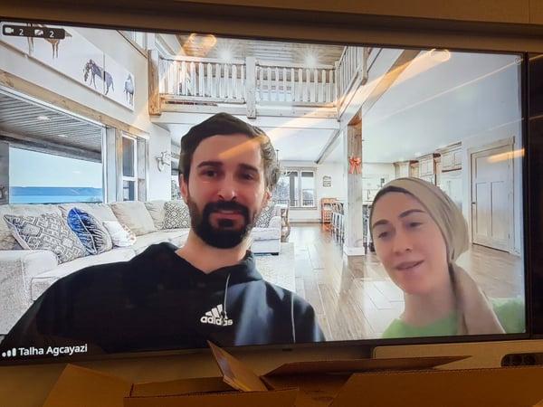 A photograph captures a large screen displaying a video conference between two people—a bearded man in a black Adidas hoodie and a woman in a beige head covering—who are using a virtual background of a luxurious, rustic interior home.