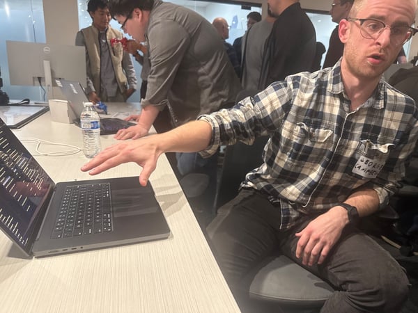A candid photograph taken indoors shows a man in glasses and a plaid shirt gesturing toward an open laptop screen, while several other people are gathered in the background around desks, suggesting a technical meeting or demo session.