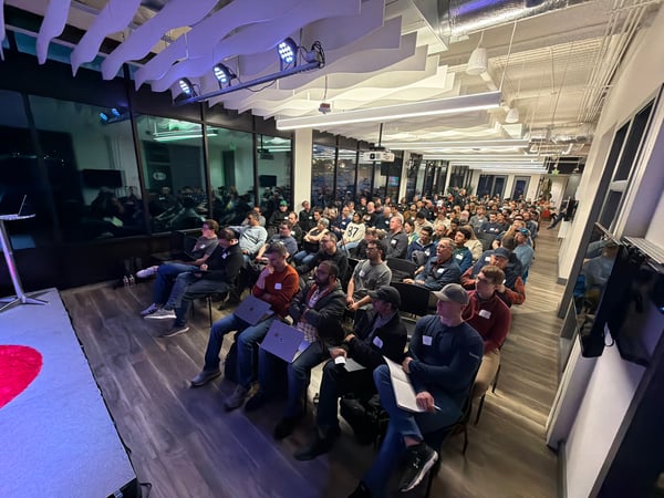 A large, predominantly male audience is seated in a modern indoor event space, facing a stage area for a presentation.