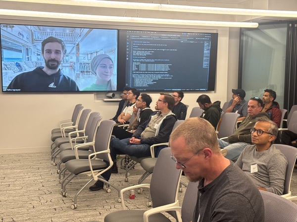 A photograph capturing an indoor technical meeting or presentation where an audience is seated, watching two large screens displaying a video conference and lines of code.