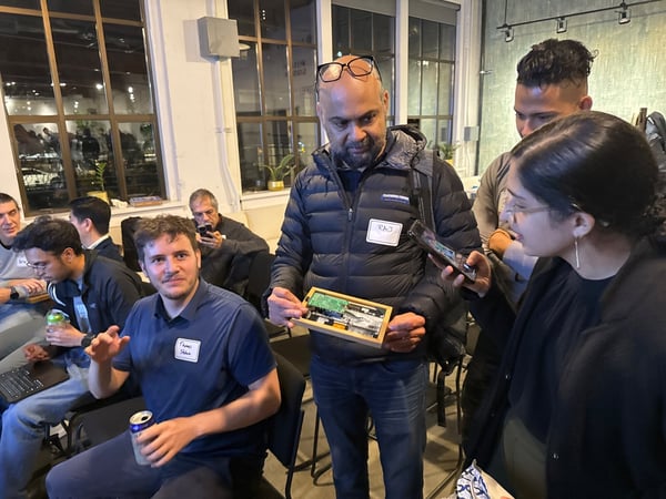 A candid indoor photograph capturing a group of people at a technical or networking event, where a man in a black puffer jacket is holding up a small electronic circuit board for others to examine. Several attendees are standing and seated in the background of the loft-style venue.