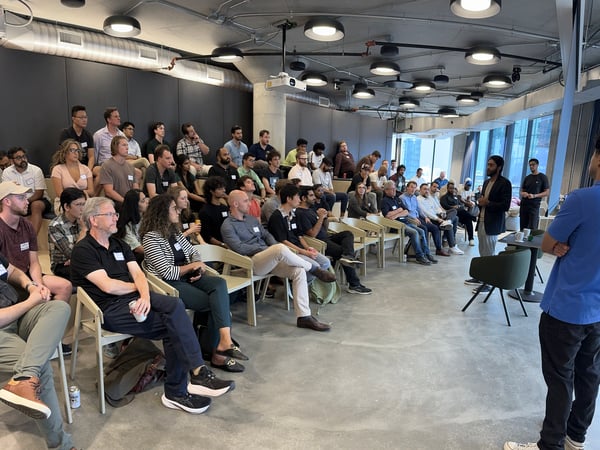 A large group of people is seated in a modern, industrial-style indoor venue, attending a presentation or seminar given by several individuals standing at the front.