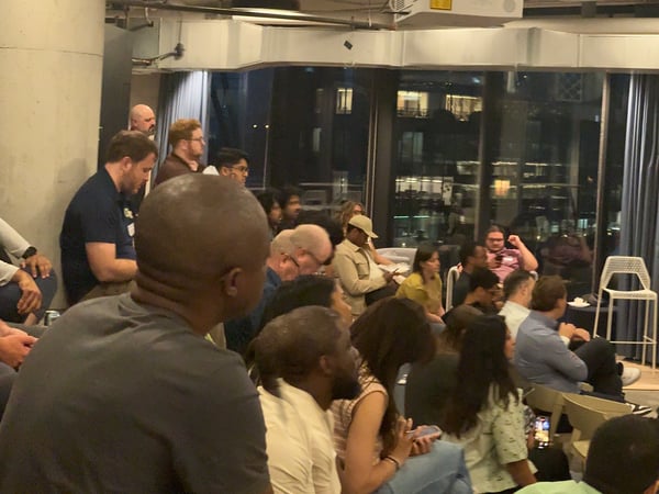 A photograph capturing a technical presentation or meetup event held indoors at night, showing a presenter addressing a large seated audience while a projector screen displays lines of code.
