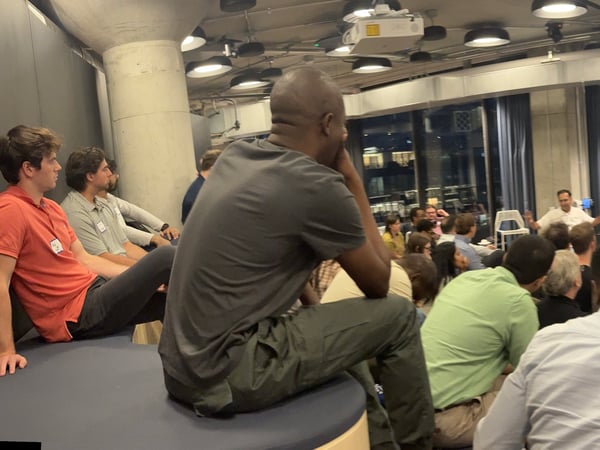 A panoramic photograph capturing an indoor technical presentation or meetup at night, showing a large audience watching a speaker and a projection screen displaying lines of code.