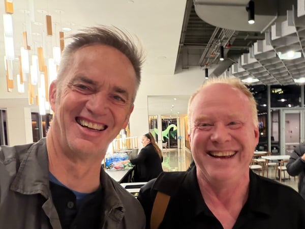 A close-up, candid selfie-style photograph of two middle-aged men smiling broadly at the camera, taken indoors in a modern venue with unique hanging light fixtures.