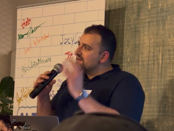 A man is giving a presentation or talk indoors, holding a microphone and gesturing, standing in front of a whiteboard covered in names and next to a screen displaying programming code.