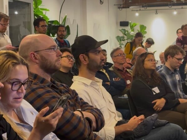 A candid photograph captures a diverse audience of approximately twenty people seated and standing indoors, all focused intently on an unseen speaker or presentation area.