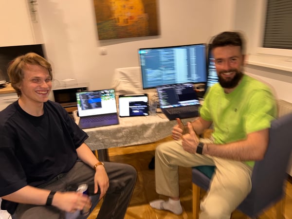 Two men are sitting at a table filled with electronic devices, including laptops and a large monitor displaying code, appearing to be working on a project together.