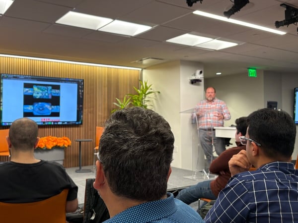 A photograph taken from the perspective of an audience member, showing a man speaking at a clear podium during an indoor presentation, with a large screen displaying technical content and a Syracuse University banner visible on the left.
