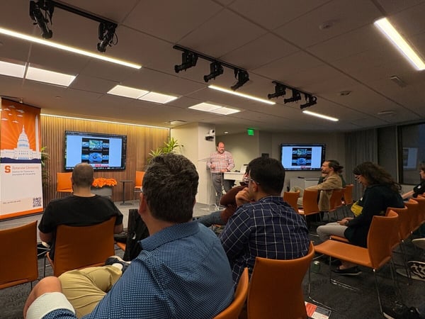 A photograph capturing an indoor presentation or lecture where a speaker addresses an audience seated in rows of orange chairs. The background features wood slat walls, two large monitors displaying data, and a prominent orange and white banner for Syracuse University.