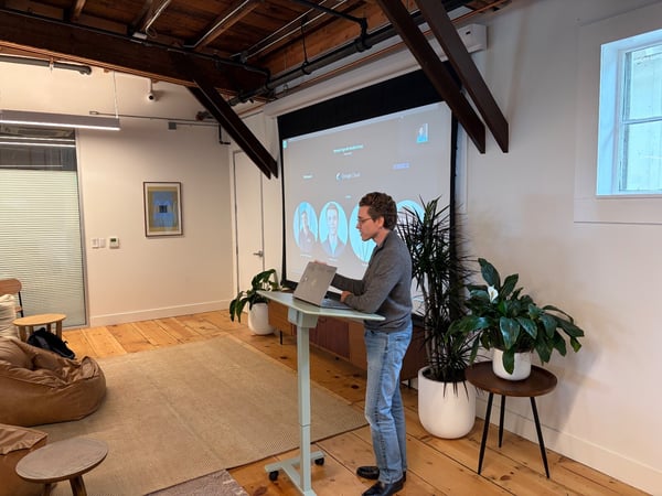 A man stands at an adjustable desk, looking at his laptop while presenting in a modern office space with exposed beams and a large projection screen displaying a video conference interface.