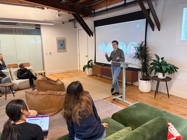 A candid photo captures a man giving a presentation in a modern, relaxed office space with exposed beams and comfortable seating, while several attendees watch, some using laptops. A large projection screen behind the speaker displays presentation content related to 'Secure Supply Chain' and 'Google Cloud'.