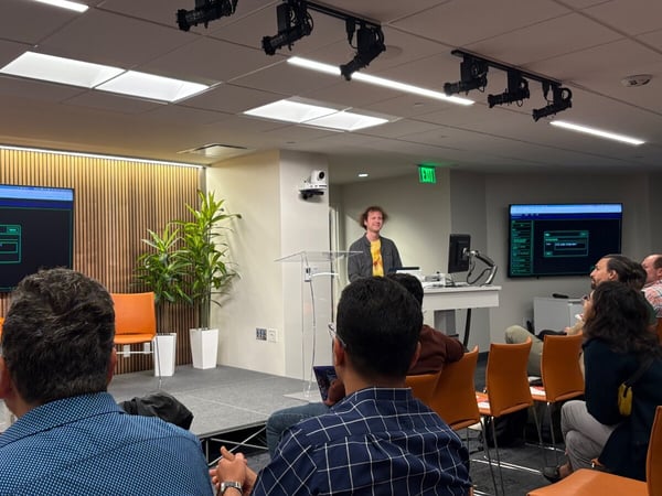 A photograph capturing an indoor technical presentation where a speaker stands on a stage behind a podium, addressing an audience seated in orange chairs. Two large monitors display dark-themed software interfaces or code on either side of the stage.