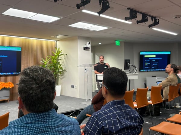 A photograph capturing an indoor technical presentation where a male speaker stands at a clear podium addressing an audience, with presentation slides displayed on large screens behind him.
