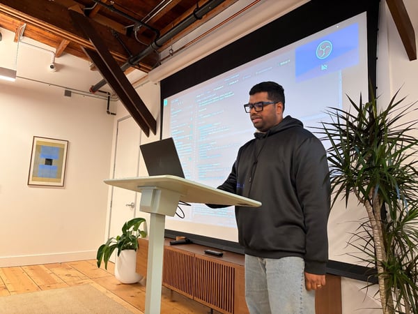 A man wearing a black hoodie and glasses stands behind a podium, giving a technical presentation in an indoor space with exposed wooden ceiling beams and a large projection screen displaying lines of code.