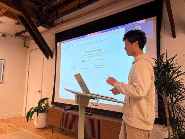 A young man wearing a light beige hoodie and pants is giving a presentation or software demonstration in an indoor setting, standing beside a large projector screen displaying a web interface related to AI coding agents.