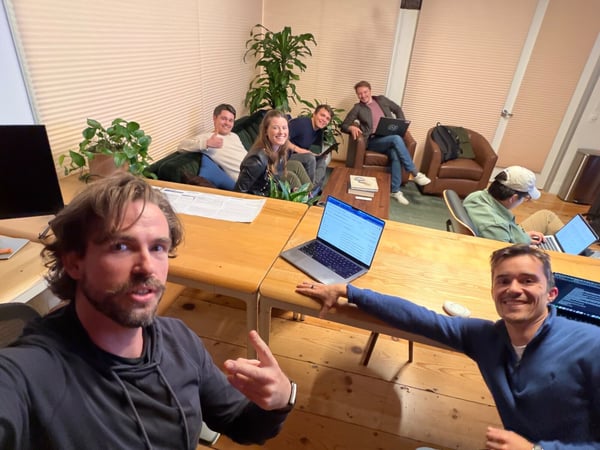 A group selfie captures six young professionals gathered in a casual, plant-filled office or co-working space, with most people smiling directly at the camera while others work on laptops.