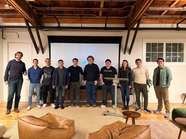 A group of ten casually dressed people stands together in a line, posing for a photo in a modern office space featuring exposed wooden ceiling beams and a large projection screen behind them.
