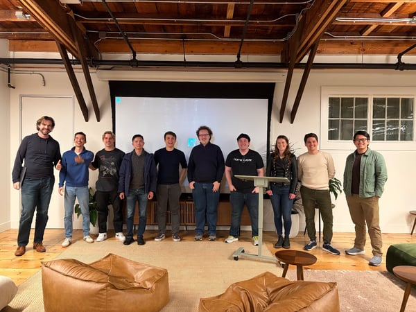 A group of ten colleagues stands in a line posing for a team photo in a modern office space characterized by exposed wooden ceiling beams and a large projection screen behind them.