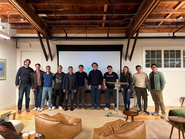 A group of eleven people, consisting of nine men and two women, pose for a photo indoors in a spacious room featuring exposed wooden ceiling beams and a large blank projection screen behind them.