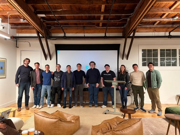 A group of eleven individuals, primarily men, pose for a team photograph indoors in a modern office space characterized by exposed wooden ceiling beams and a large, mostly white projection screen behind them.