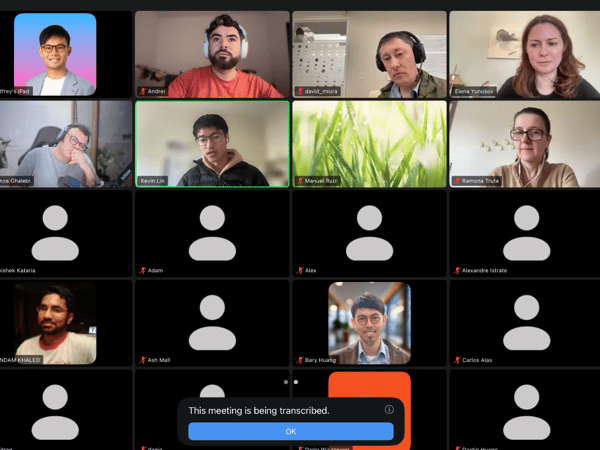 A screenshot of a video conference call displayed in a grid layout, featuring several participants visible via live video or profile pictures, interspersed with many tiles showing only default black screens and names. A notification overlay at the bottom indicates that the meeting is being transcribed.