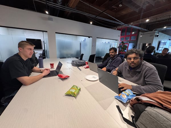 Three men are sitting around a large, light-colored conference table with open laptops, bags of chips, and drinks, appearing to be engaged in a work session or collaboration in a modern office environment.