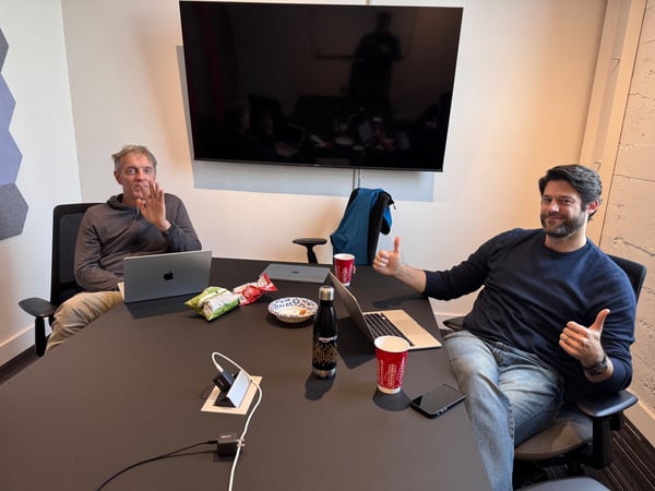 A candid photo capturing two men seated at a dark conference table in an office setting, surrounded by laptops, snacks, and drinks; the man on the right is smiling and giving two thumbs up.
