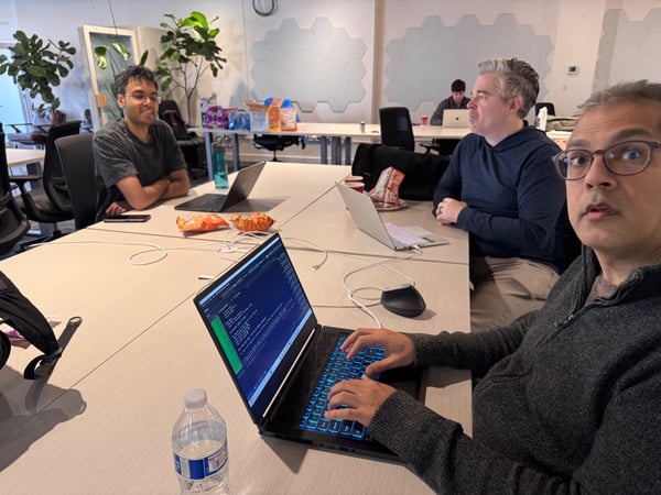 A candid photograph captures four men gathered around a large table in a modern office, with the man closest to the camera typing on a laptop displaying code.
