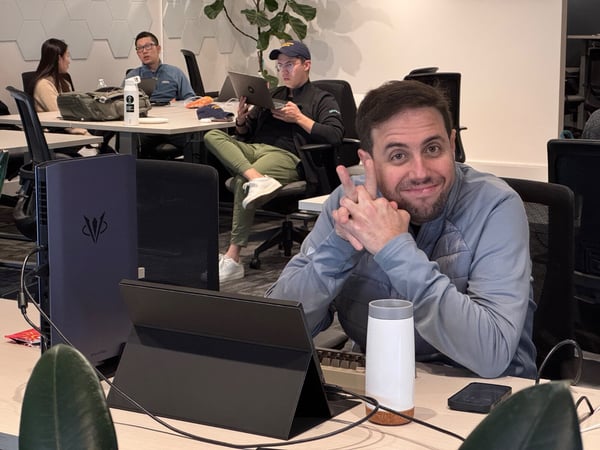 A candid photograph taken in a modern office space shows a man in the foreground smiling directly at the camera, while three colleagues work or converse at desks behind him.